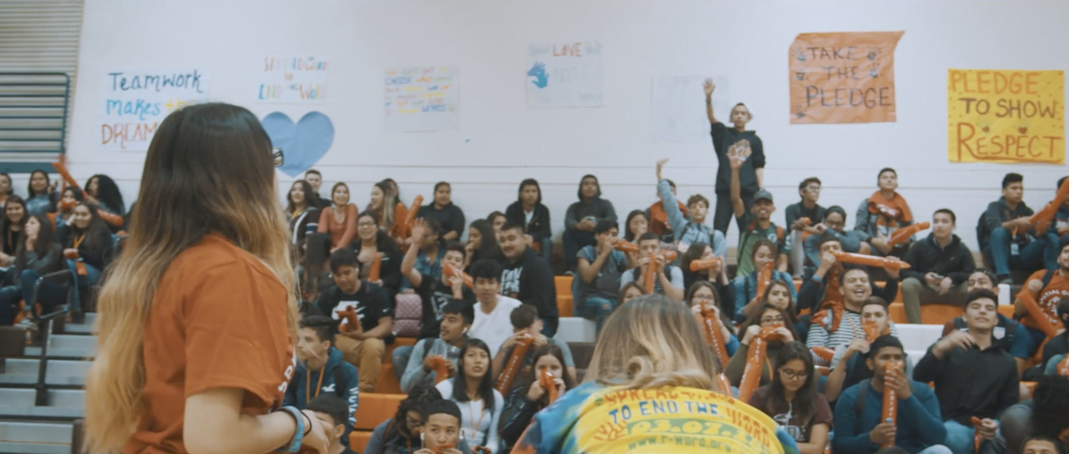 Students sitting in the bleachers of an assembly as two students face them.