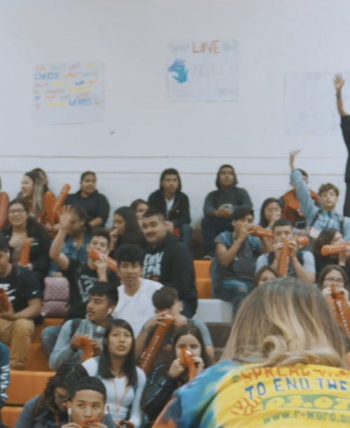 Students sitting in the bleachers of an assembly as two students face them.