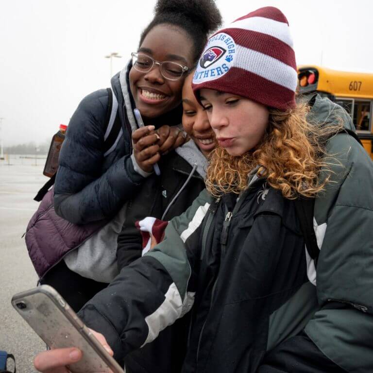 Three girls posing for a selfie. They are wearing winter gear and are standing in front of a school bus.