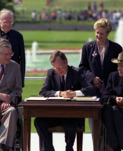 President George Bush signs into law the Americans with Disabilities Act of 1990 on the South Lawn of the White House. L to R, sitting: Evan Kemp, Chairman, Equal Employment Opportunity Commission, Justin Dart, Chairman, President's Committee on Employment of People with Disabilities. L to R, standing: Rev. Harold Wilke and Swift Parrino, Chairperson, National Council on Disability, 07/26/1990.