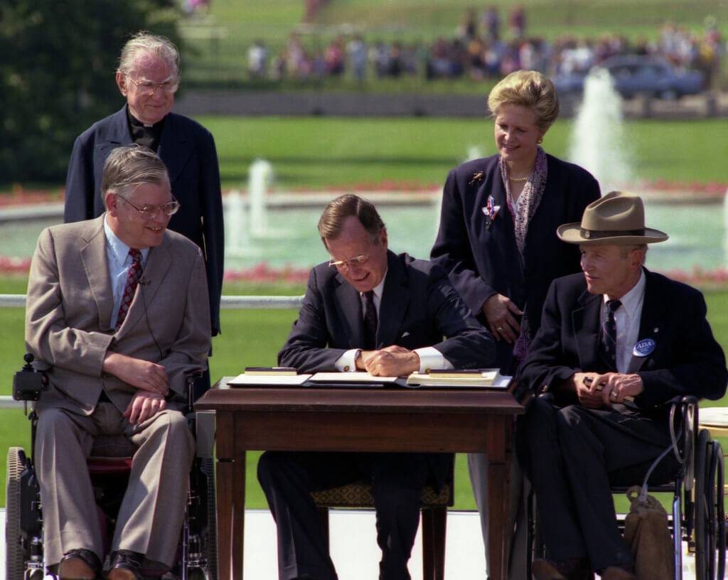 President George Bush signs into law the Americans with Disabilities Act of 1990 on the South Lawn of the White House. L to R, sitting: Evan Kemp, Chairman, Equal Employment Opportunity Commission, Justin Dart, Chairman, President's Committee on Employment of People with Disabilities. L to R, standing: Rev. Harold Wilke and Swift Parrino, Chairperson, National Council on Disability, 07/26/1990.