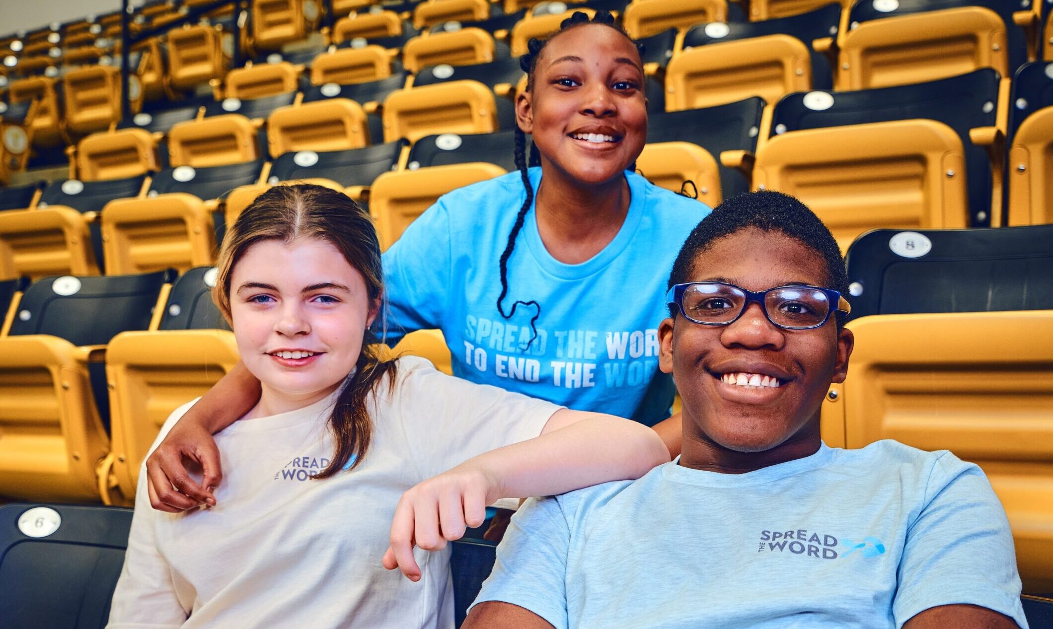A trio of young individuals in Spread the Word tshirts sitting in a stadium.