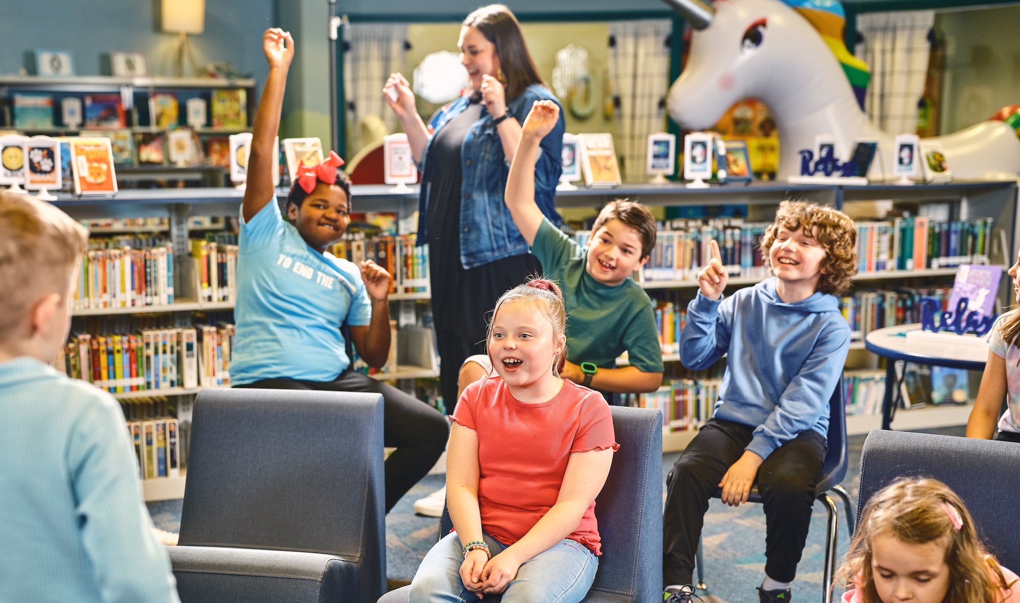 Children in a library eagerly raise their hands, participating in a group activity or discussion.