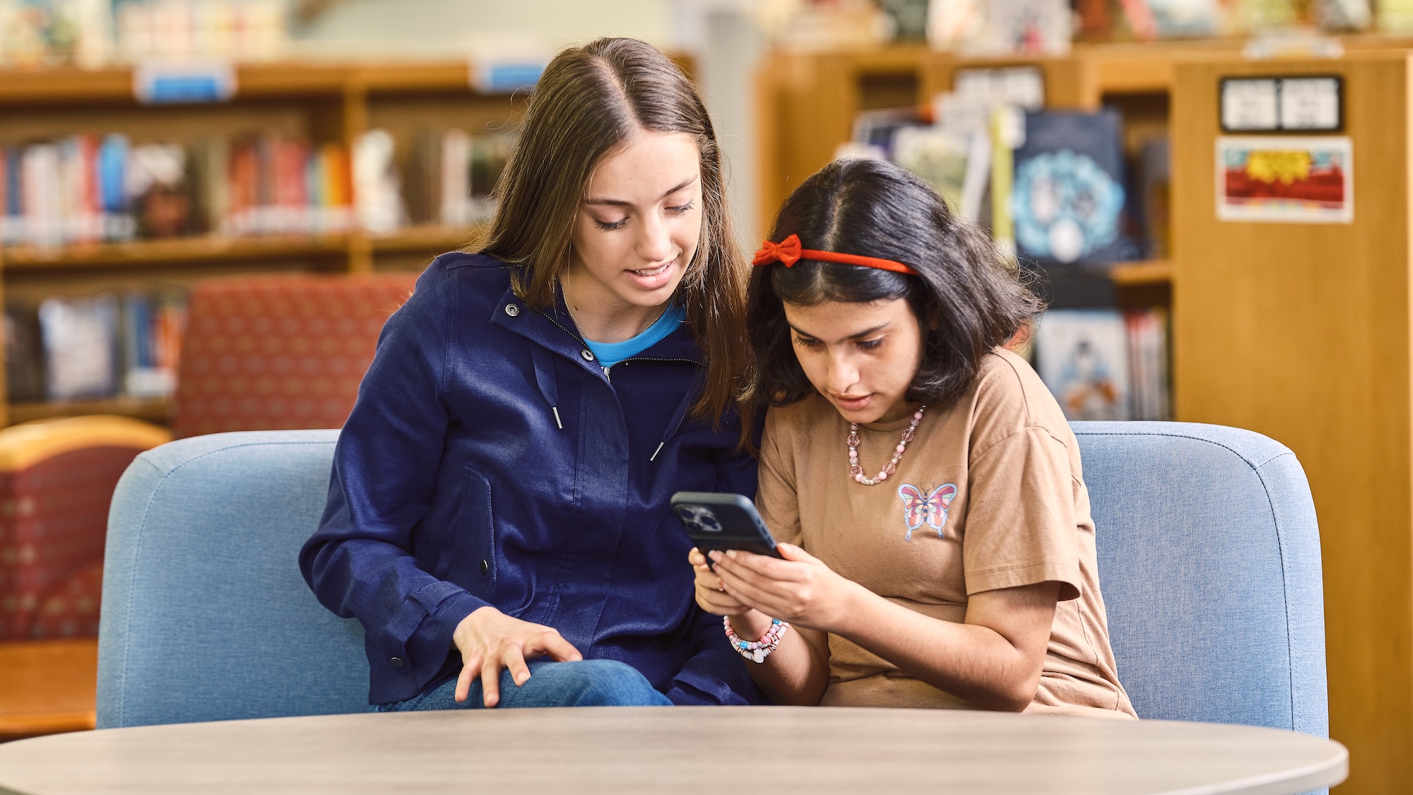 Two girls sitting on a couch in a library, focused on a cell phone together.